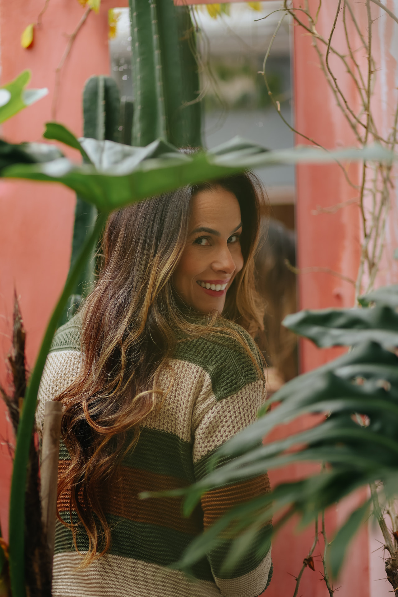 Kamilla Flor sorrindo, vista entre folhagens, diante de uma parede rosada